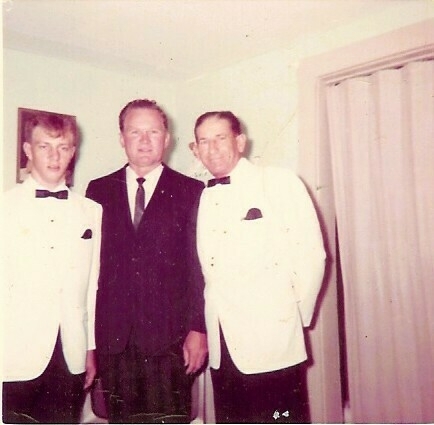 Auto-generated description: Three men pose together indoors, with two wearing white tuxedos and the third in a black suit.