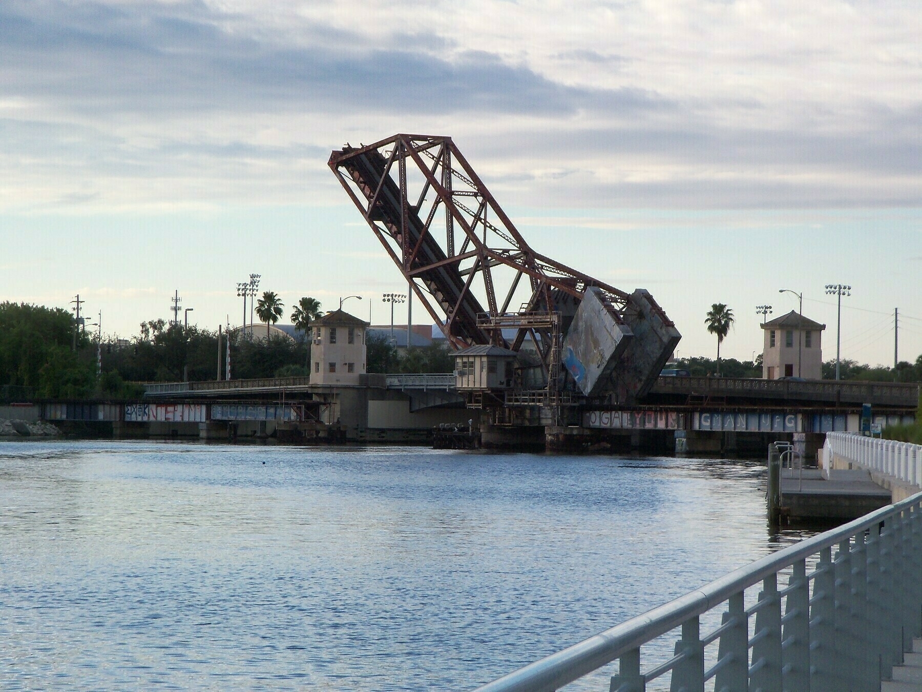 A drawbridge is partially raised over a body of water with a cloudy sky in the background.